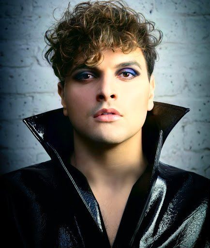 A man with curly hair and blue eyeshadow is wearing a black jacket