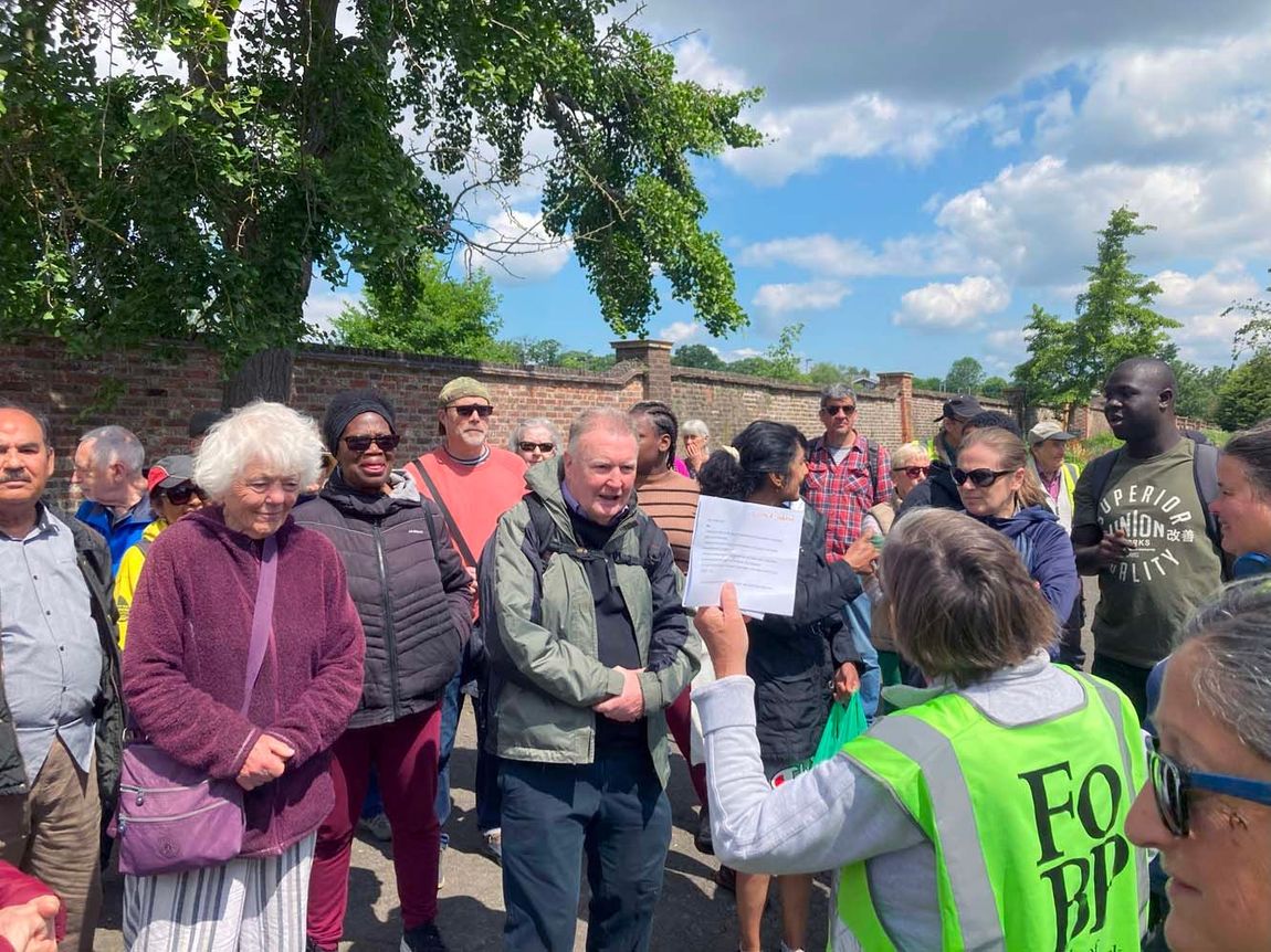 People gathered outdoors at a protest, one person in a green vest addressing the crowd.