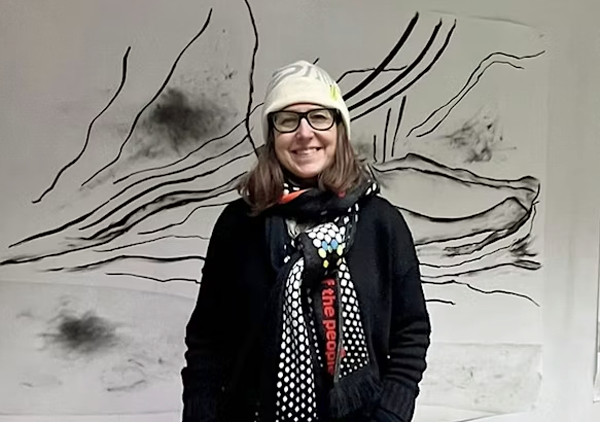 Smiling person in glasses and white hat standing before abstract line artwork on a wall