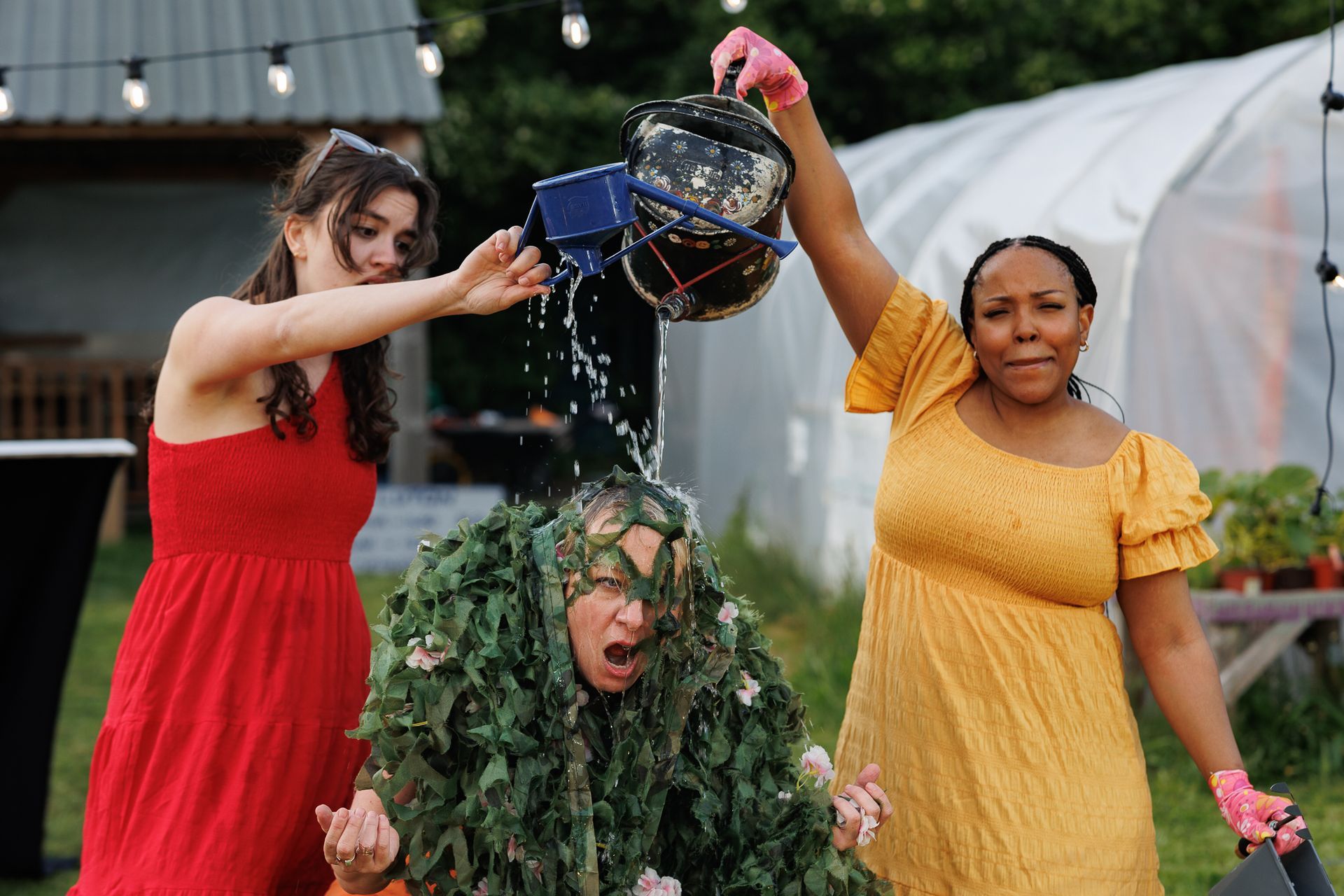 Three people outdoors pour water from a bucket onto a person in a leafy costume, all wet and smiling