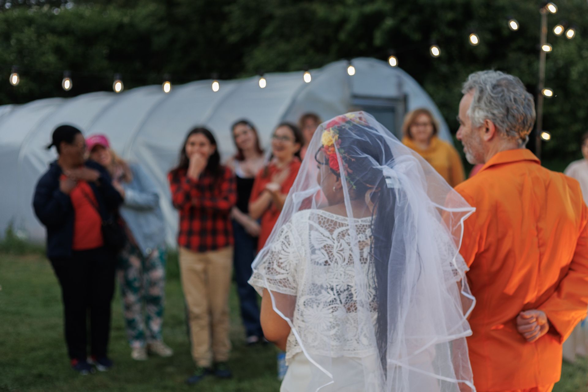 Bride in white veil stands with an older man in an orange shirt at an outdoor evening gathering with guests and string lights