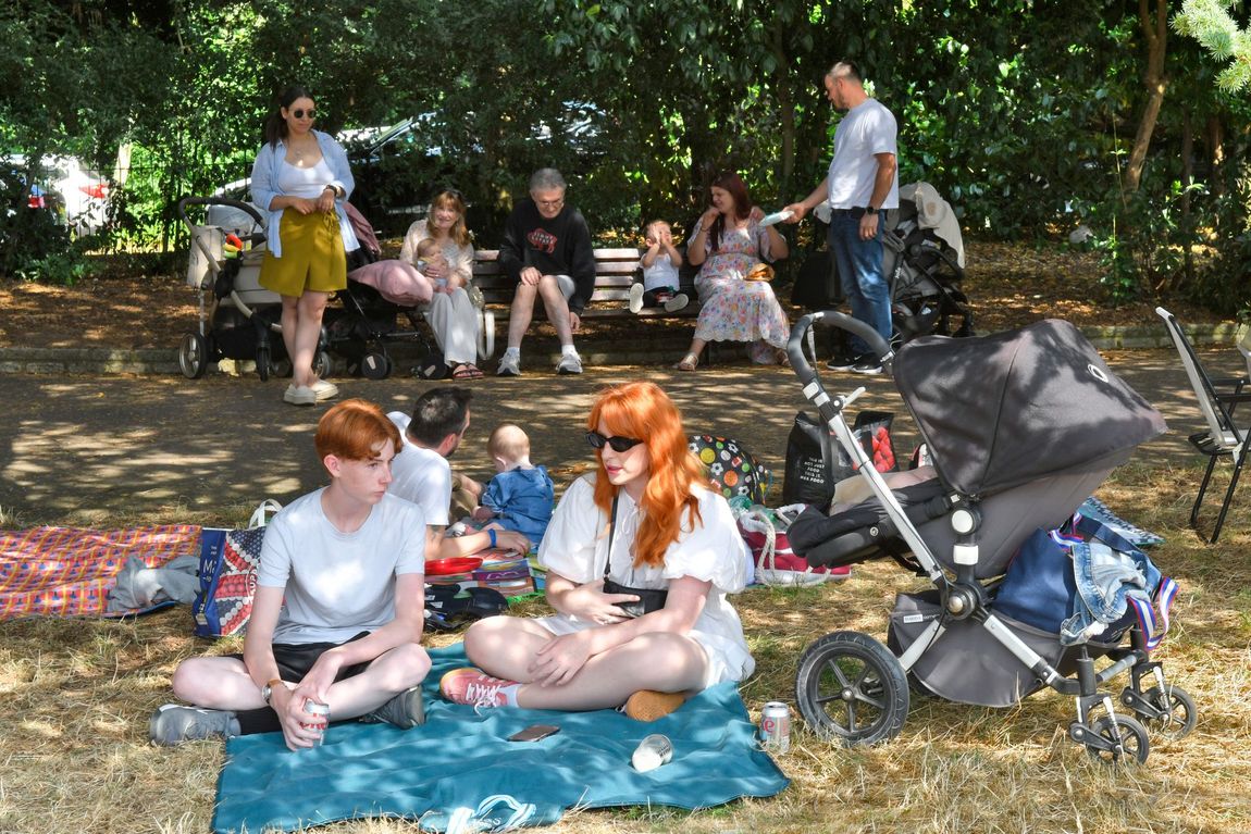 People resting on blankets in a shady park, with a stroller and picnic gear nearby.