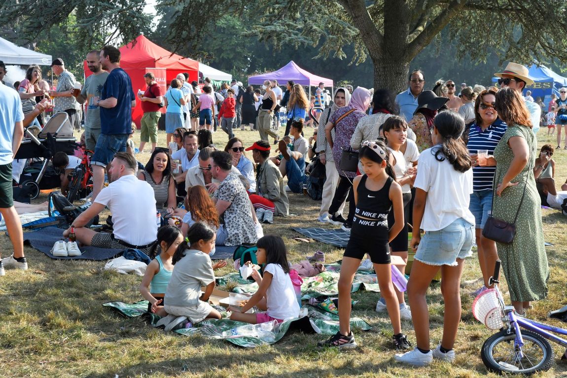 Crowd of people relaxing at an outdoor park gathering with tents, trees, and bicycles on a sunny day