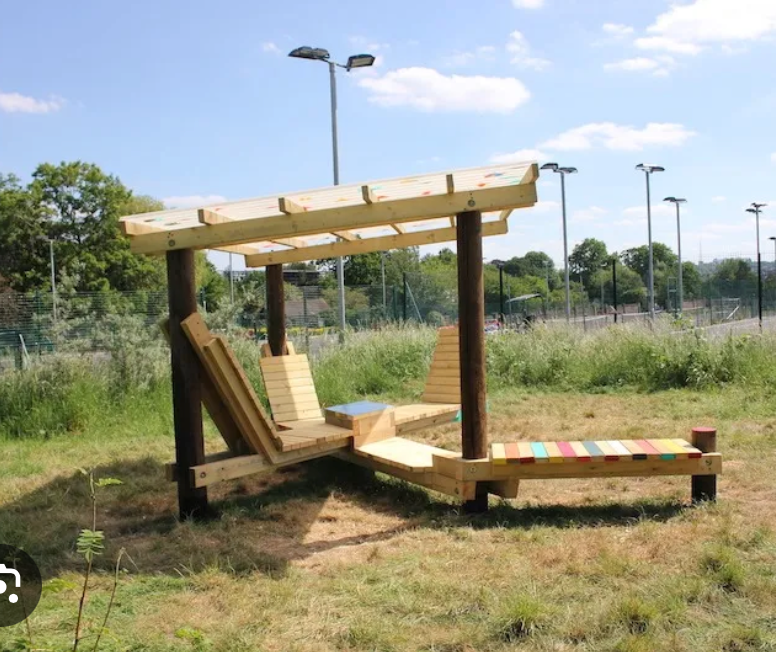 Wooden outdoor lounge bench with canopy in a grassy park, with tall light poles in the background