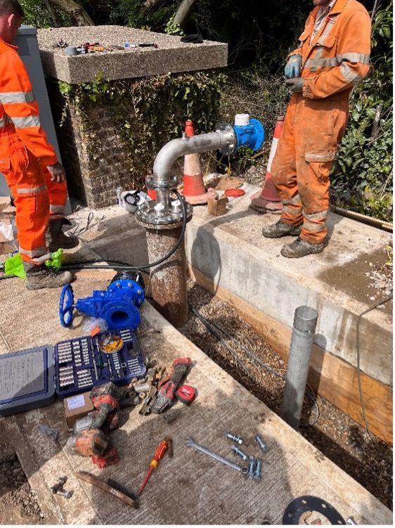 Workers repairing a blue water valve and pipe at a roadside utility pit with tools scattered nearby