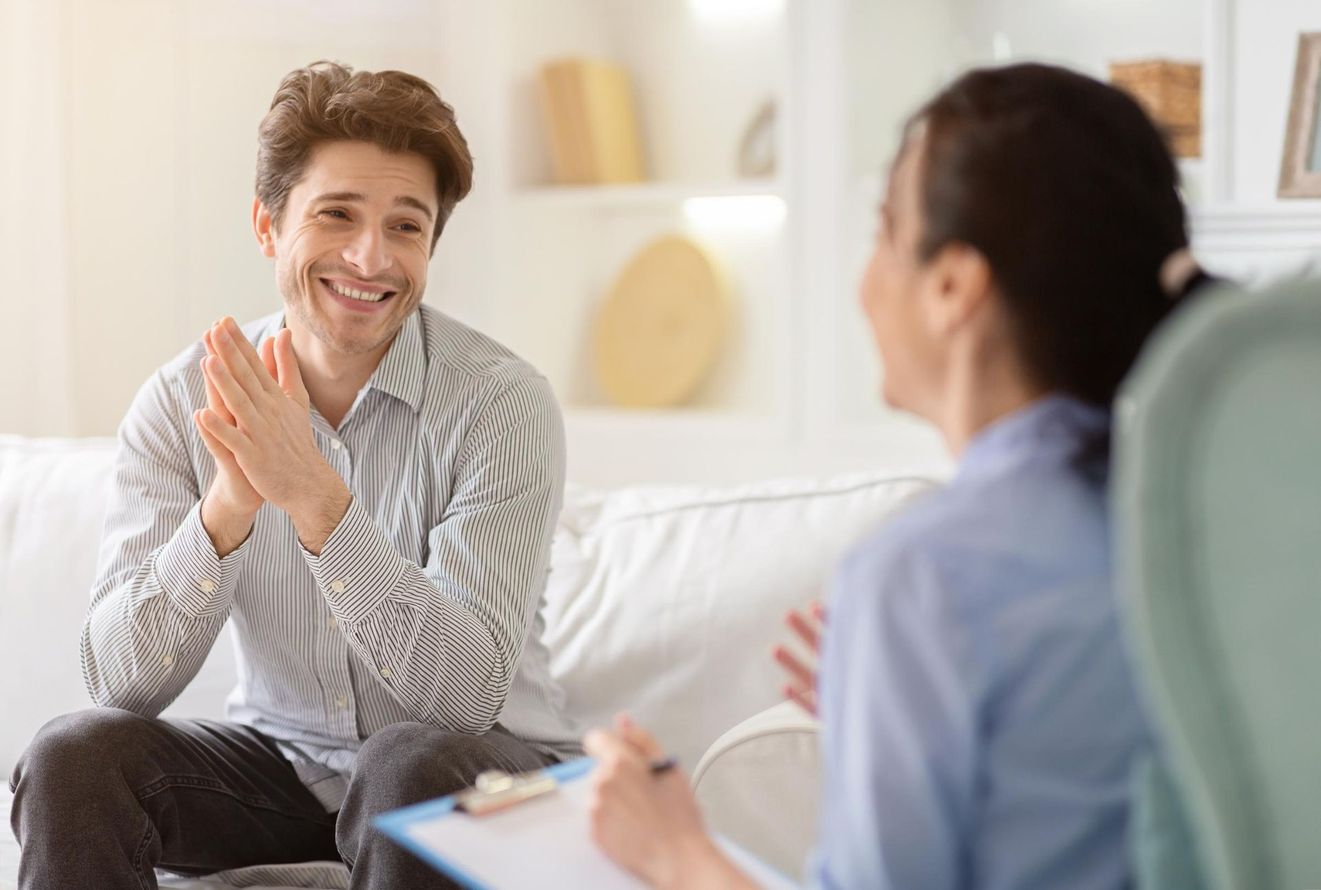 A man is sitting on a couch talking to a woman who is holding a clipboard.