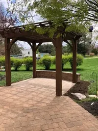 Brown wooden pergola on a brick patio with a low curved brick wall. Green lawn and bushes in the background.