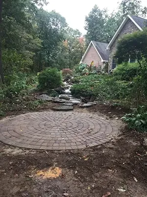 Circular brick patio in front of a natural stone stream leading to a house in a wooded area.