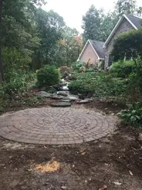 Circular brick patio in front of a natural stone stream leading to a house in a wooded area.