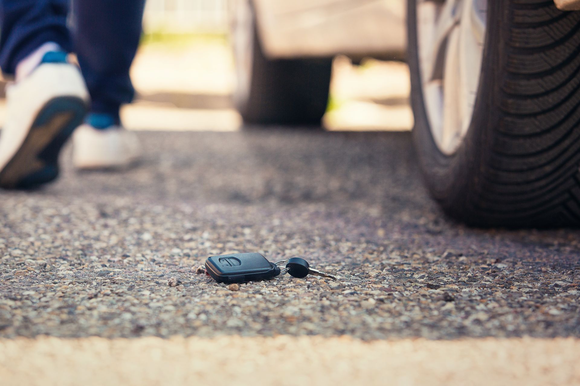 Car keys lying on asphalt near a car's tire; person walking away in the background.
