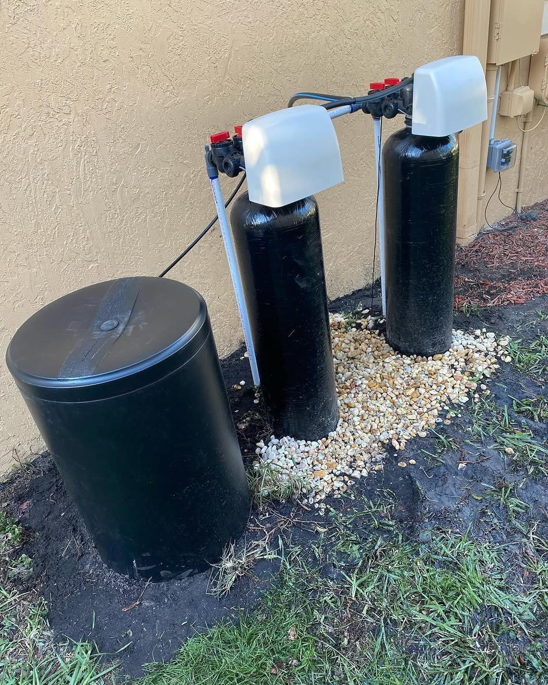 Two black water softeners are sitting in the dirt in front of a house.