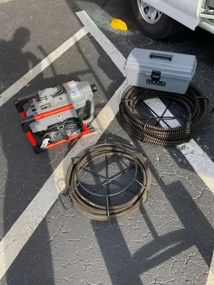 Drain cleaning equipment: electric auger, toolbox, and two coiled drain snakes on pavement.