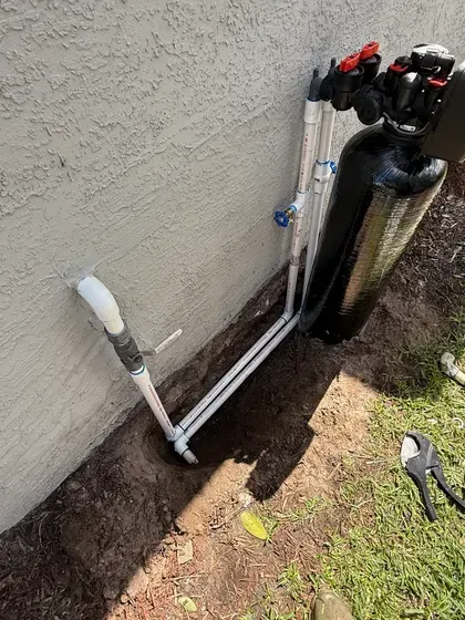 Pipes and a black water filter system are installed along a concrete wall near grass, with a trench visible.