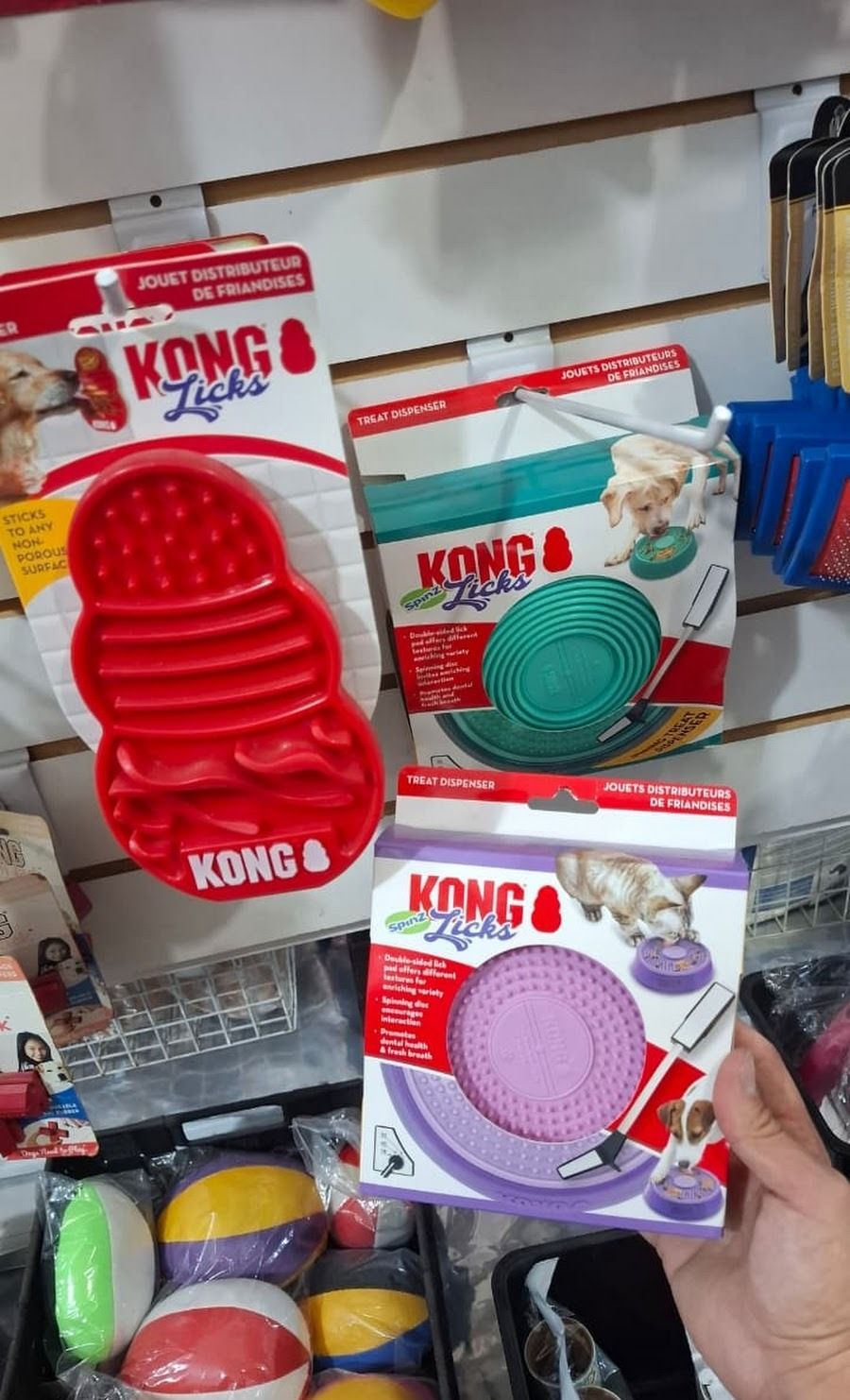 Una mano sostiene cajas de juguetes para perros Kong de color rojo, turquesa y morado en un estante de una tienda.
