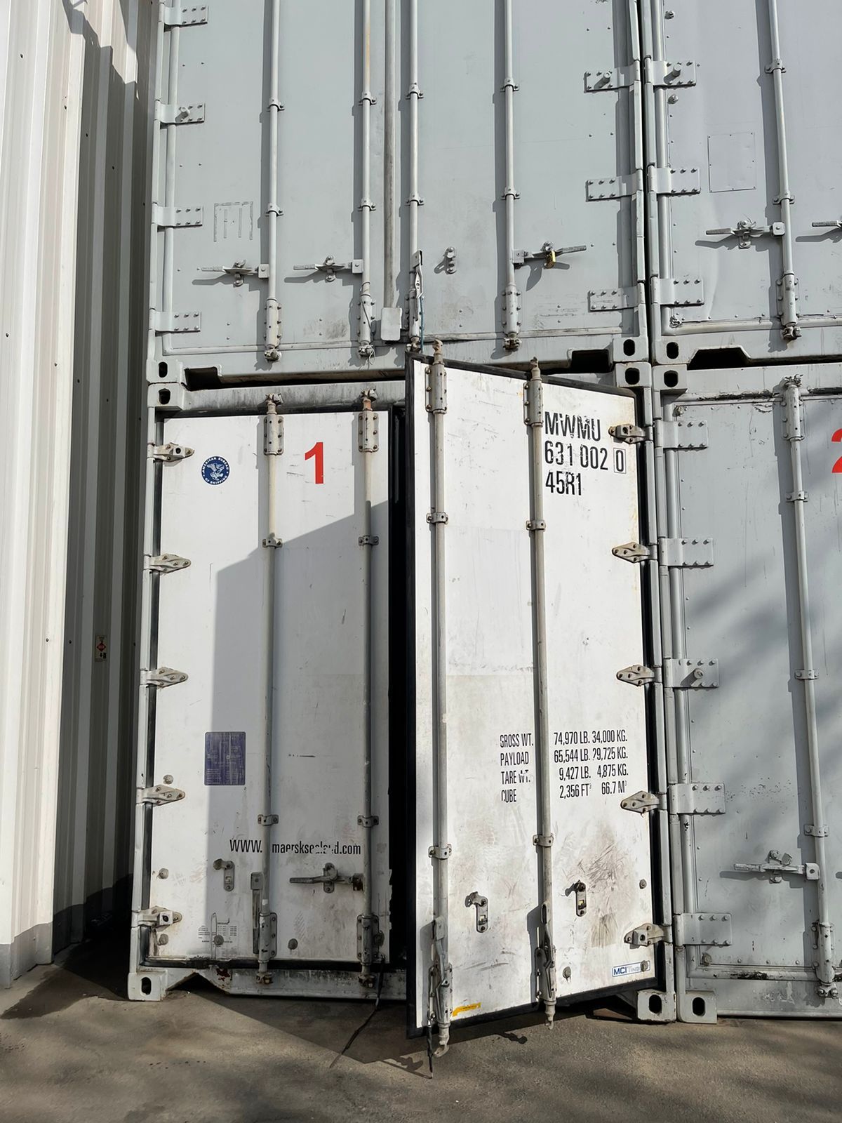 Fila de contenedores de transporte blancos en una zona de carga industrial cubierta, con un pequeño quiosco a la derecha.