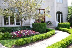 A curved stone walkway leads to a grand entrance framed by manicured hedges, a colorful flower bed, and a leafy tree.