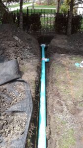 A blue PVC pipe with a T-junction laid in an open dirt trench in a backyard.