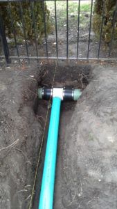 A blue PVC pipe T-junction connected to an existing underground pipe with black rubber couplings in an outdoor trench.