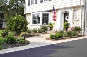A stone walkway leads to the front entrance of a house, surrounded by well-manicured bushes, flowers, and an American flag.