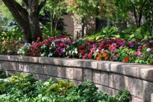 A stone retaining wall in front of a garden filled with vibrant, colorful flowers and lush green trees.