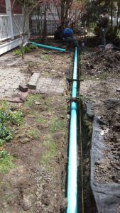 A trench in a yard containing a long blue pipe, with a worker in the background and a house on the left.