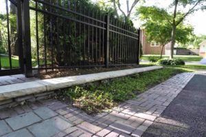 A black metal fence runs behind a stone retaining wall bordering a paved brick walkway and asphalt driveway.