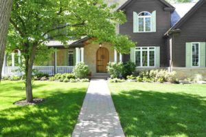 A two-story dark brown house with stone siding, light green shutters, and a walkway leading to a wooden front door.