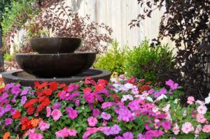 A tiered dark stone fountain surrounded by vibrant red, pink, and purple flowers in a garden setting.