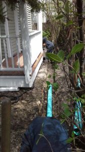 A person works in a garden next to a house porch, with blue drainage pipes running along the ground nearby.