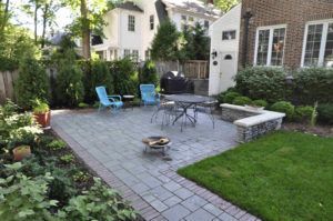 A stone patio in a backyard with a dining set, two blue chairs, a small fire pit, and a low stone wall, bordered by a lawn.