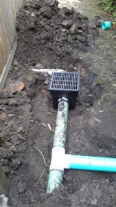A black plastic catch basin connected to PVC drainage pipes in a dirt trench next to a wooden fence.