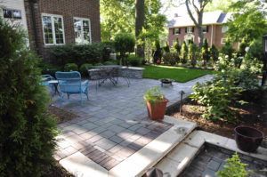 A patio with grey stone pavers, blue outdoor chairs, and a small dining set, situated next to a brick house and garden.