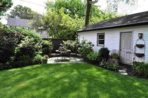 A lush green backyard featuring a small stone patio with a bistro table, a white shed, and surrounding trees and shrubs.