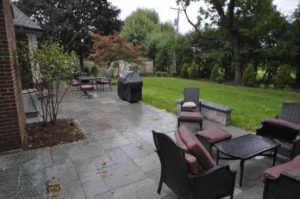 A stone patio with outdoor seating, a grill, and a lush green lawn in a backyard setting.