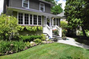 A two-story grey house with a white front porch, surrounded by lush green landscaping and trees under a sunny sky.