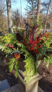 A decorative winter planter filled with greenery, red berry branches, and pinecones, sitting on a stone pedestal outdoors.