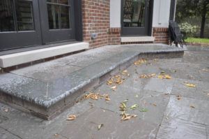 A stone patio features a low built-in bench leading to double doors on a brick house, with scattered fallen leaves.