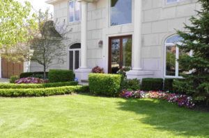 A large, multi-story house with a beige facade, white pillars, a manicured lawn, and colorful garden beds.