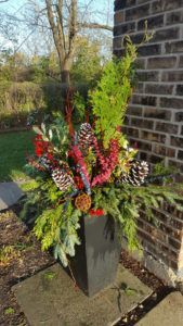 A tall, dark square planter sits on a patio with a winter arrangement of evergreen boughs, red berries, and pinecones.