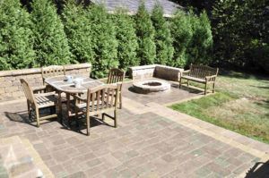 A stone patio featuring a wooden dining set, a circular fire pit, and a wooden bench, set before a row of tall green trees.