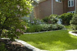 A vibrant green lawn surrounded by stone-bordered flower beds, lush foliage, and a brick house in the background.