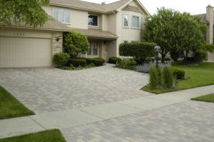 A suburban two-story house with a beige exterior and a patterned stone paver driveway.
