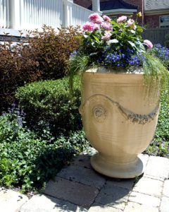 A large, light-colored ceramic planter filled with pink and blue flowers sits on a stone patio in a garden setting.