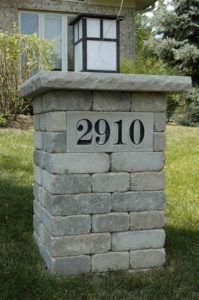A gray stone pillar displaying the address number 2910, topped with a lamp, standing on a residential lawn.