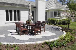 A patio with a dark wood table and chairs on a gravel base next to a white brick house with a landscaped garden.