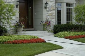 A stone house entrance with a concrete path, a lush lawn, red flower borders, and potted plants by the front door.