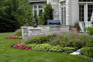 A stone patio area with a built-in grill, framed by lush landscaping featuring purple flowers and red foliage.
