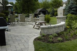 A paved stone patio with dining furniture, a small wooden picnic table, and a curved decorative stone seating wall.