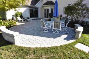 A circular stone patio features a table set with chairs, a blue umbrella, and curved stone seating walls near a house.