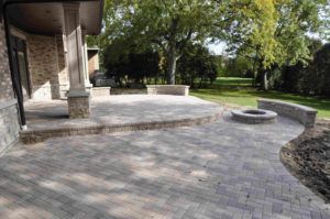 A stone patio with a raised platform, a circular fire pit, and curved stone seating walls in a backyard setting.